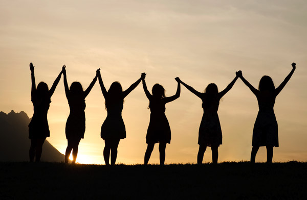 Six women are silhouetted in the sunset; their arms are raised and they are holding hands in jubilation.
